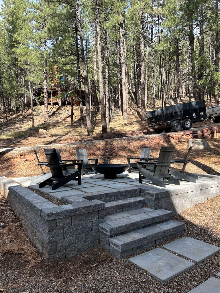 Outdoor fire pit area with modern chairs on a raised paver patio surrounded by forest trees.