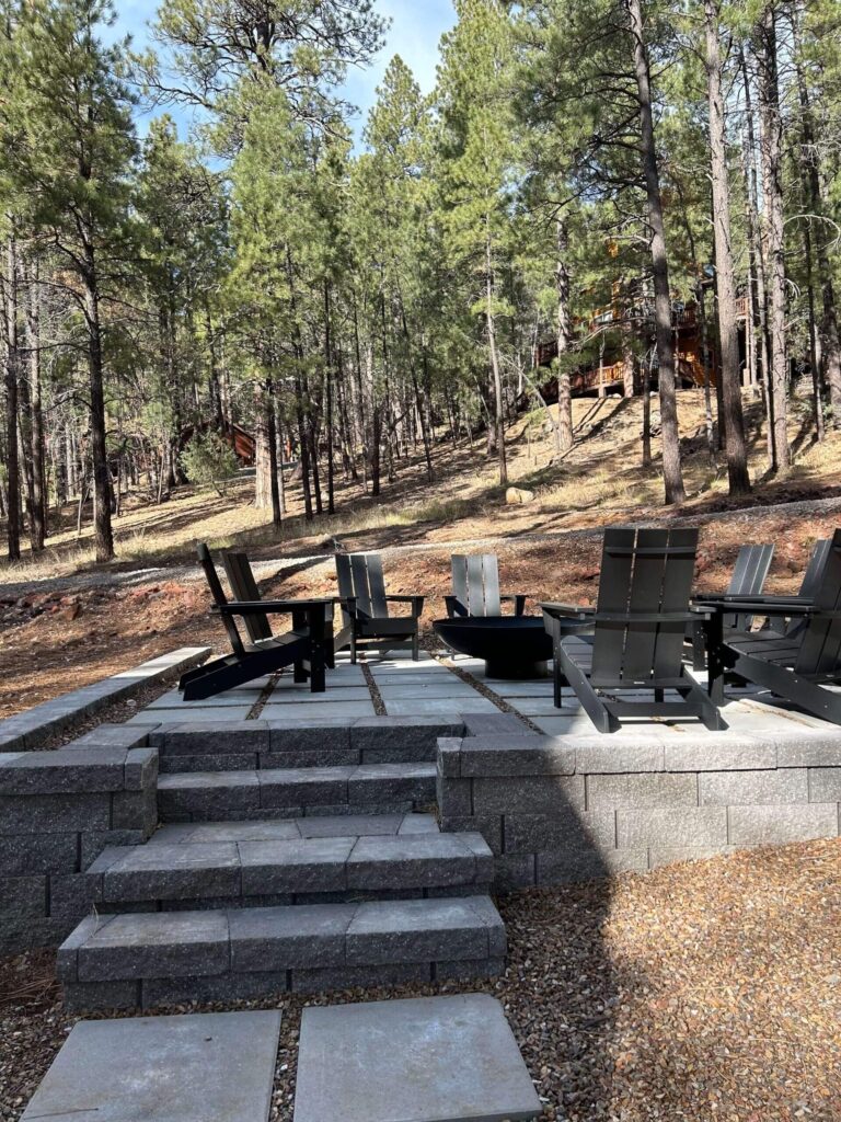 Outdoor fire pit area with modern chairs on a raised paver patio surrounded by forest trees.