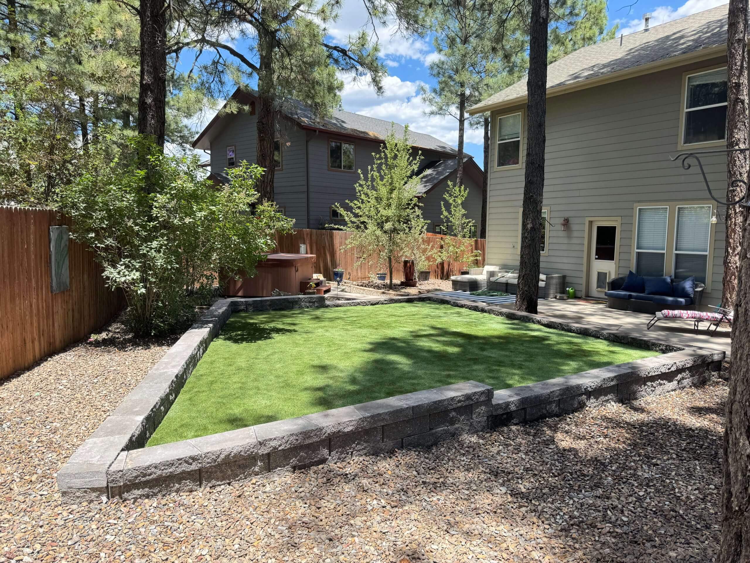 Backyard with artificial turf bordered by stone retaining walls, surrounded by gravel and mature trees.