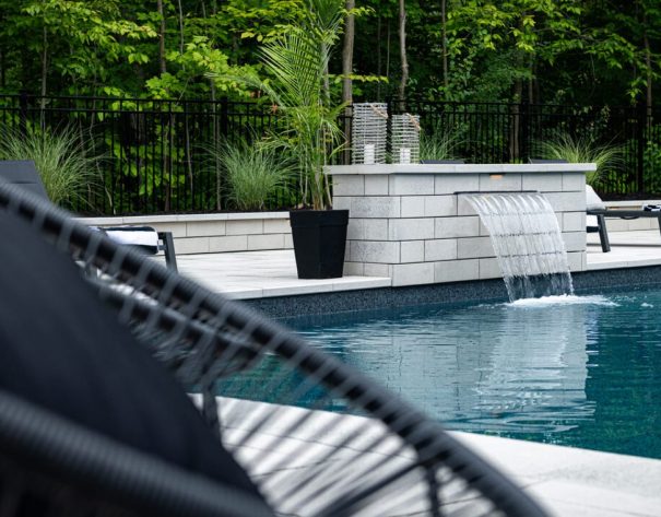 Modern backyard pool with elegant stone waterfall feature, surrounded by lush greenery and sleek lounge chairs.